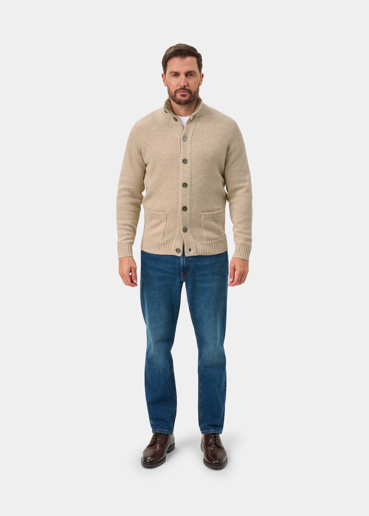 A man wears the Landford Lambswool Buttoned Mock Neck Jumper in Linen with blue jeans and brown shoes on white.