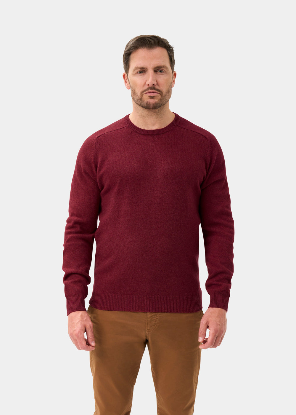 Man in Lenzie Lambswool Crew Neck Jumper in Garnet, regular fit, and brown pants stands against a light background.