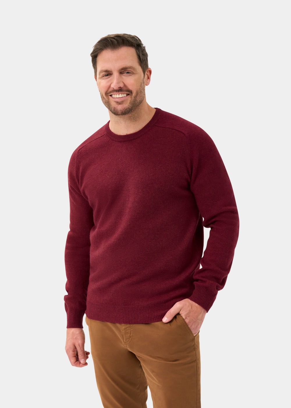 Smiling man wears Lenzie Lambswool Crew Neck Jumper in Garnet, regular fit, and brown pants against a plain background.