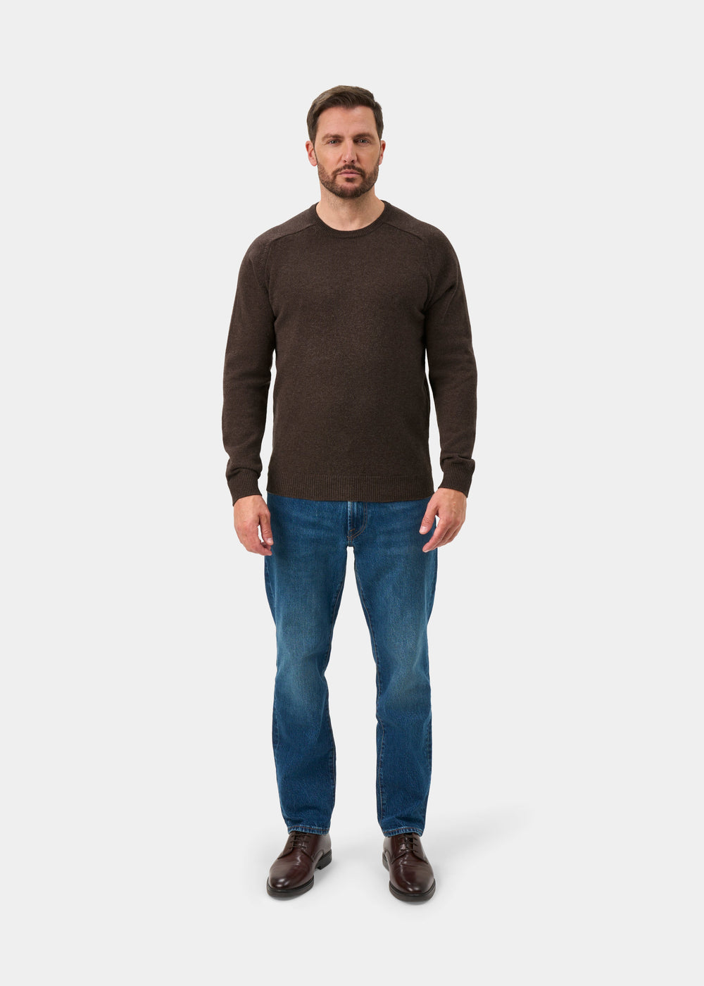 Man wearing the Lenzie Lambswool Crew Neck Jumper in mocha with blue jeans and brown shoes, plain background.