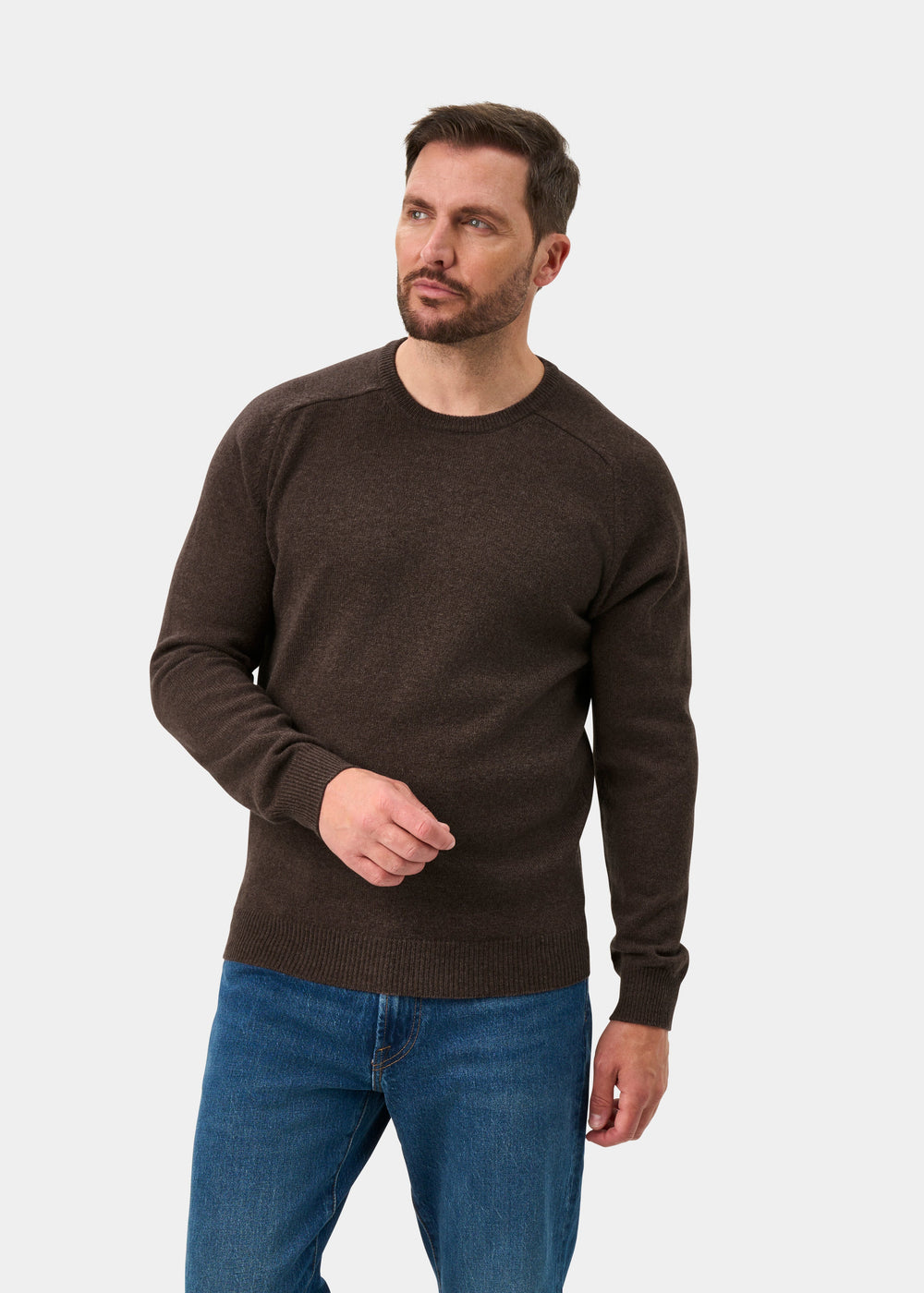 Man in Lenzie Lambswool Crew Neck Jumper in Mocha, regular fit, with blue jeans, stands against a plain light background.