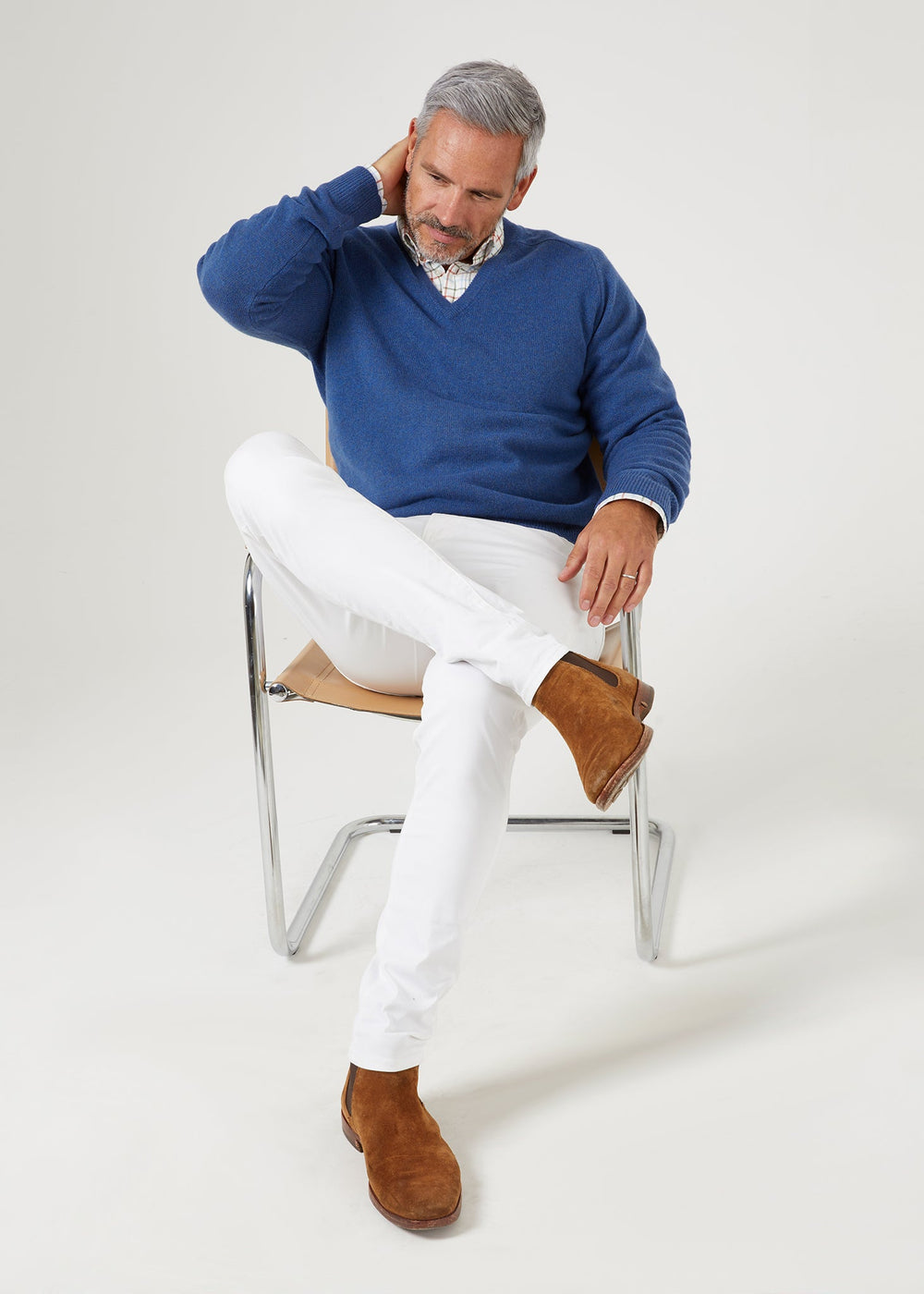 A man in a Selkirk Pure Cashmere V-Neck Jumper In Denim, plaid shirt, and white trousers sitting on a chair.