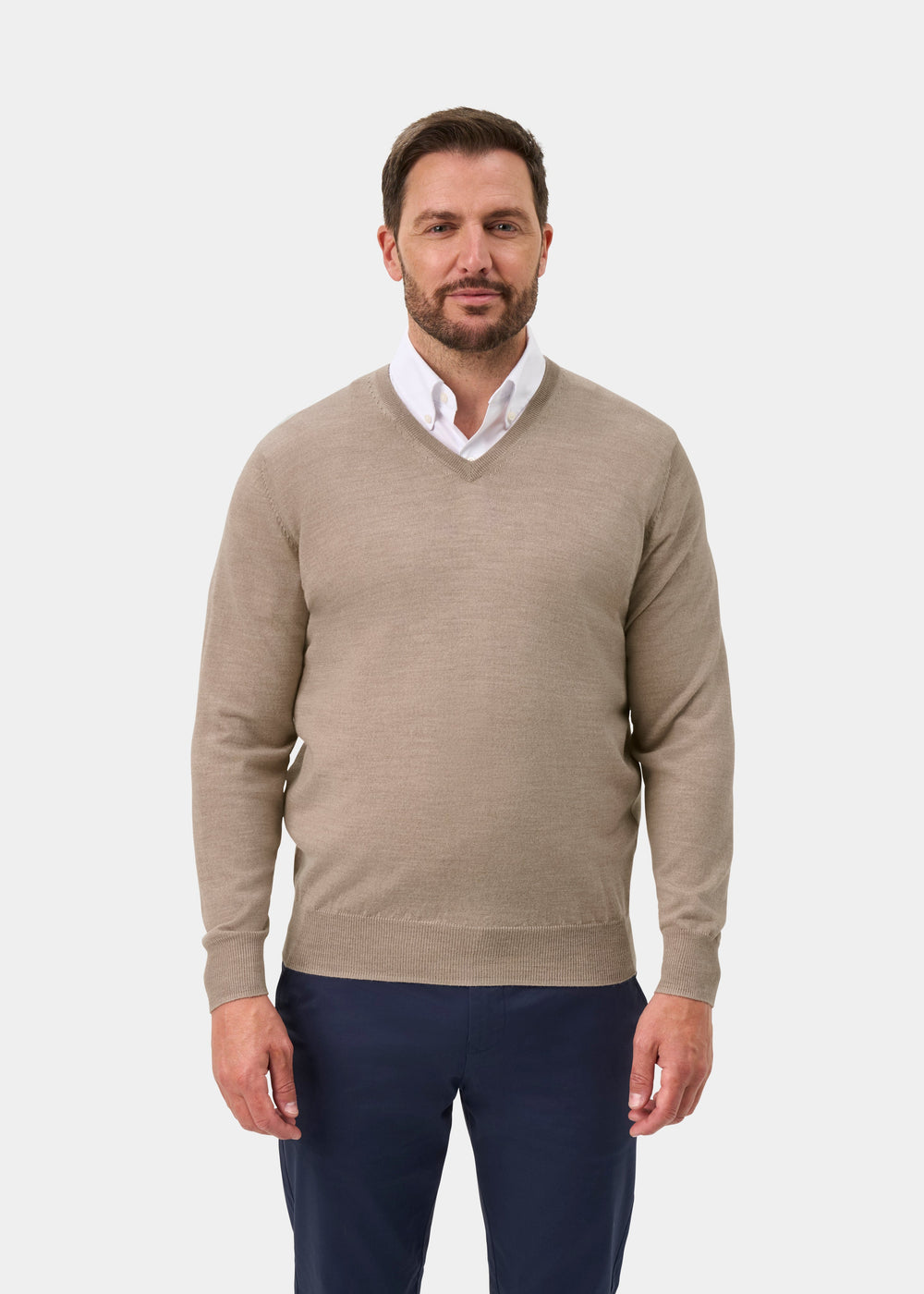 A man wearing a beige v-neck knit sweater over a white collared shirt and navy trousers, standing against a light gray studio background.
