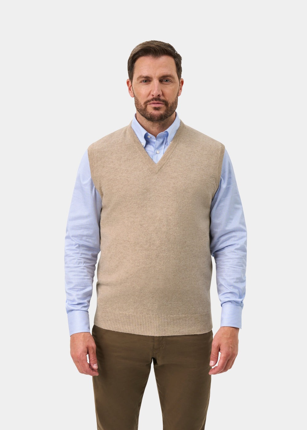 A man wearing a beige knit sweater vest over a light blue collared shirt and brown trousers, standing against a light gray studio background.