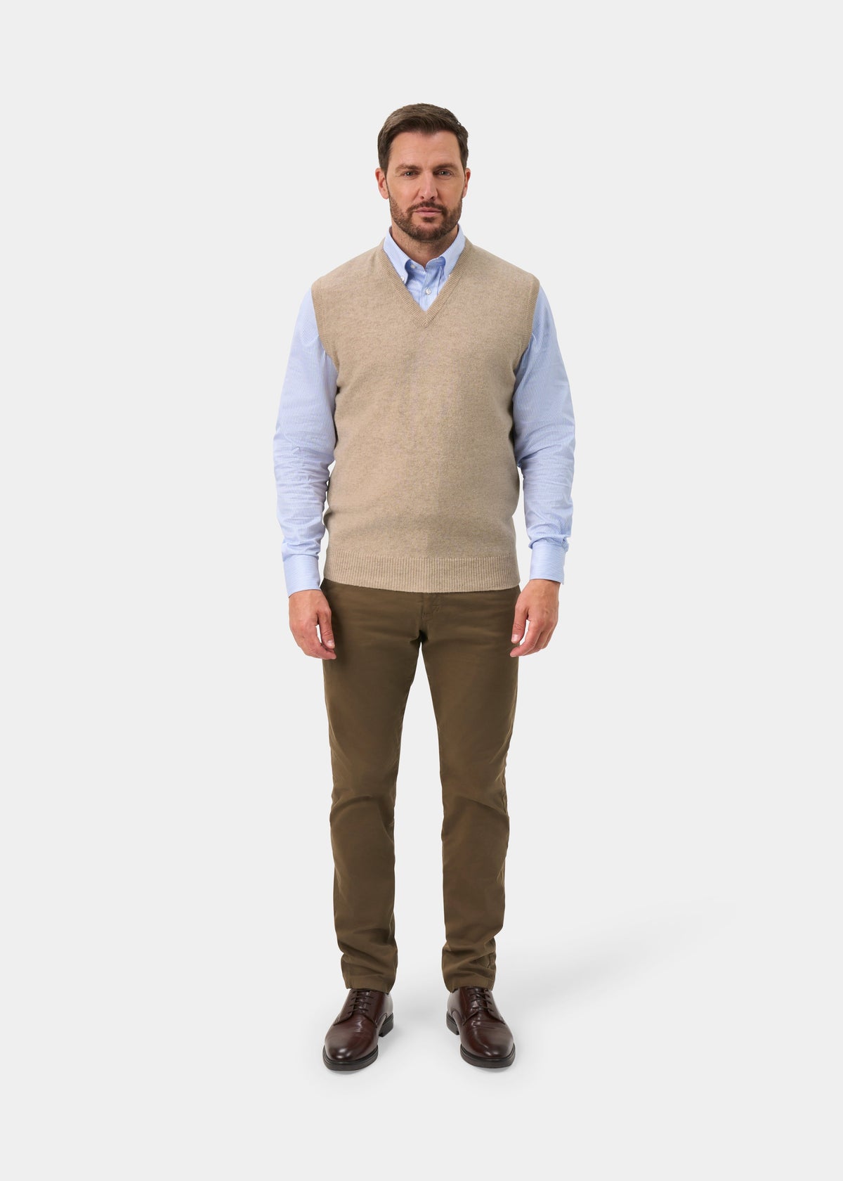 A man modeling a beige v-neck knit sweater vest over a light blue dress shirt, paired with olive brown trousers and dark brown leather shoes, against a light gray studio background.