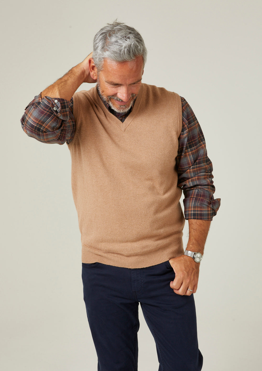 A man wearing a Norfolk Lambswool V-Neck Slipover In Camel over a plaid shirt and dark trousers.